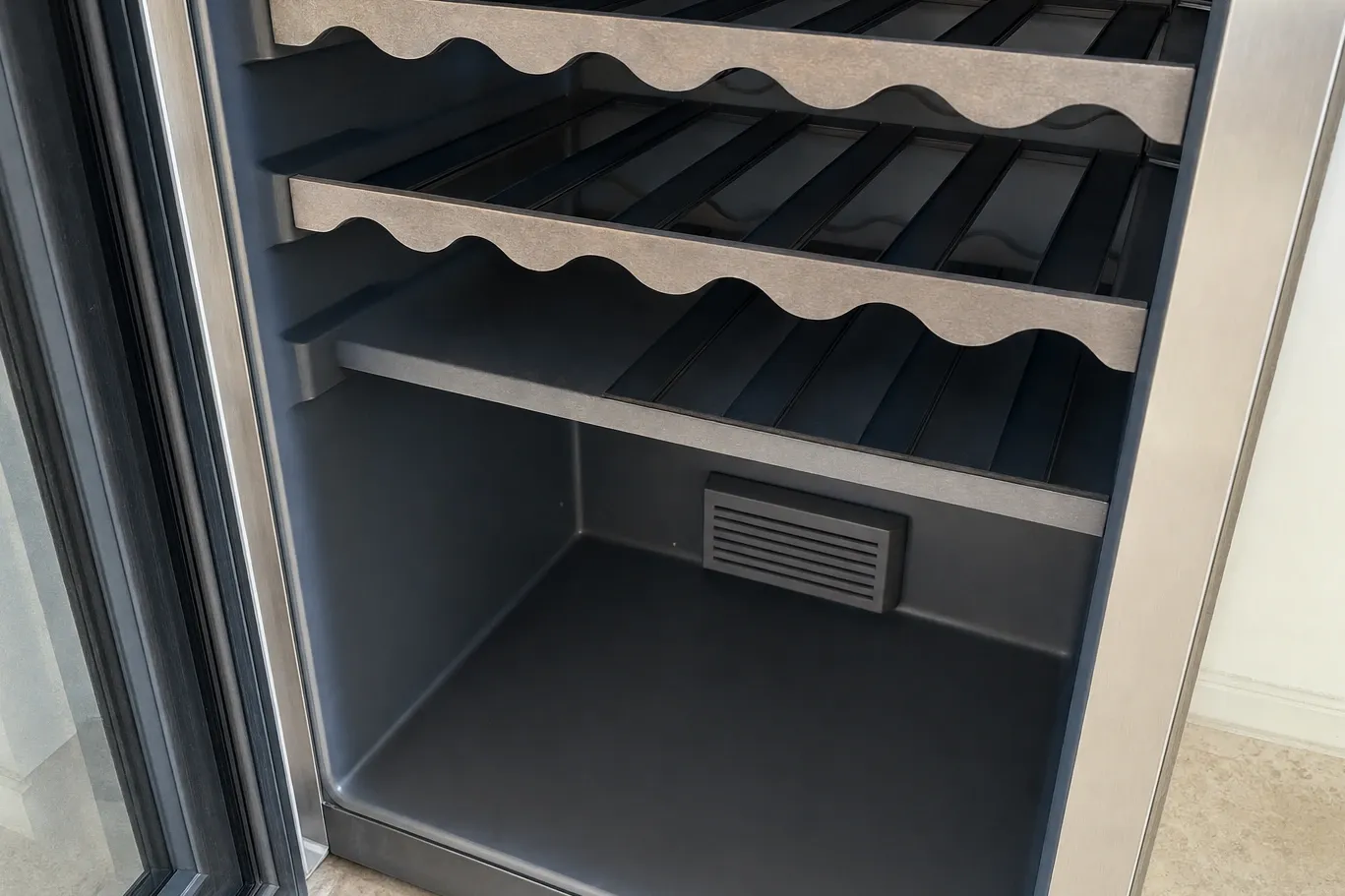 Interior of an empty wine cooler: three scalloped wooden racks, dark liner and a louvered vent low on the rear wall