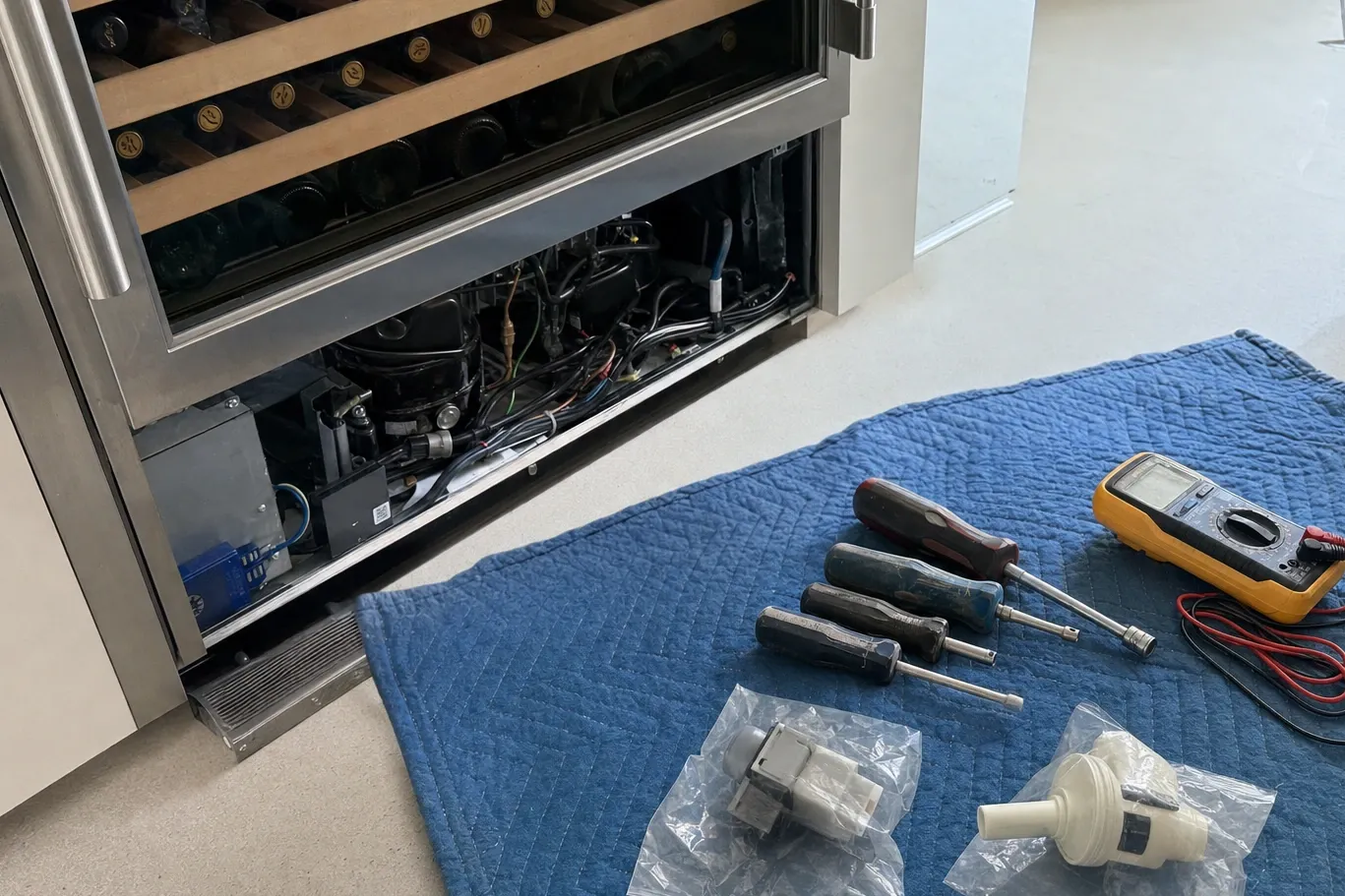 Wine column full of bottles on wooden racks with the lower grille off, compressor bay open and tools laid out on a blanket