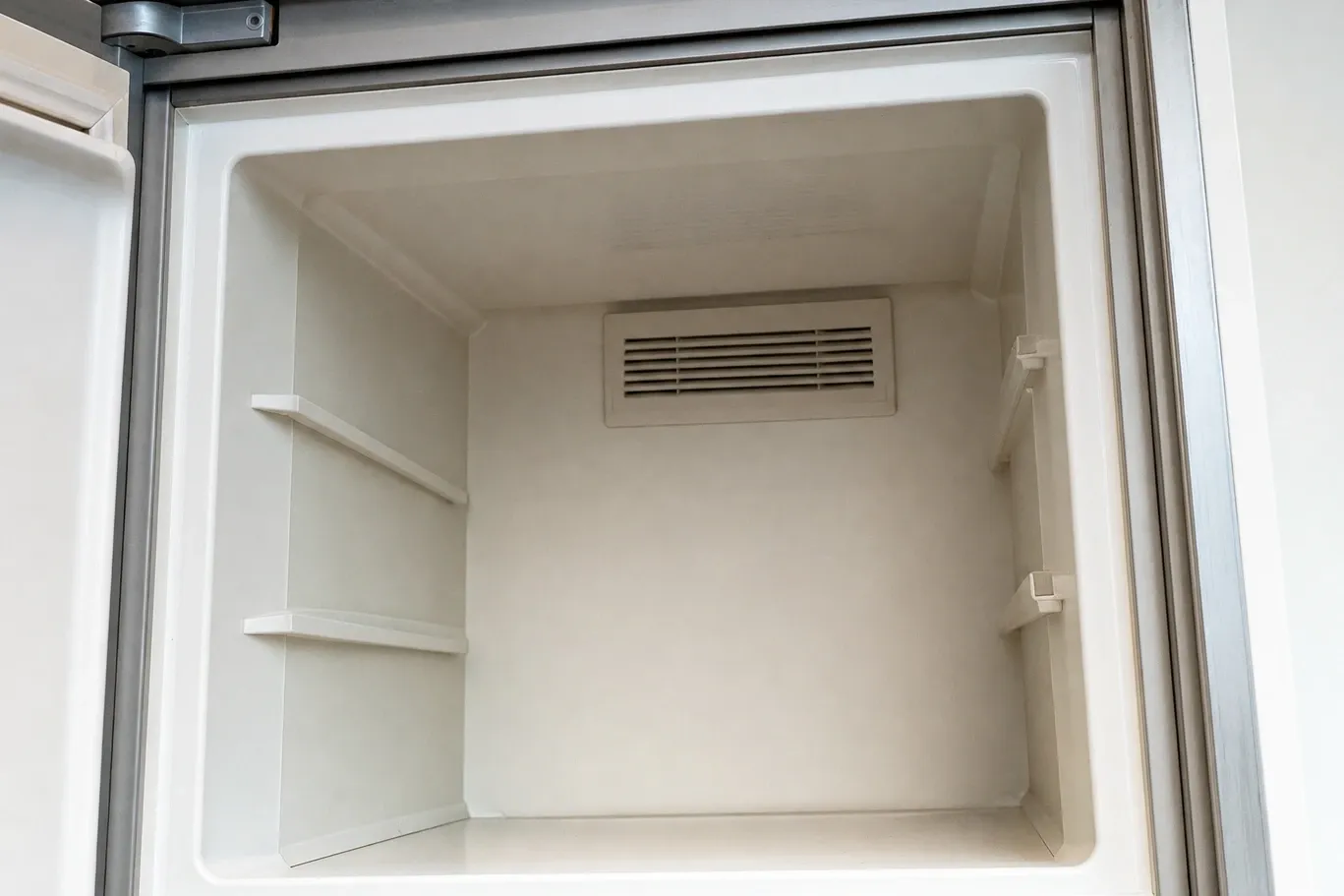 Empty ivory compartment of a built-in with the louvered air duct in the centre of the rear wall and both shelf ledges bare